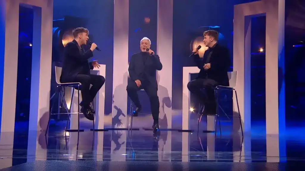 “THE MOMENT THAT STOPPED TIME…” 😢 — SIR TOM JONES BROUGHT AN ENTIRE NATION TO TEARS….At 84, Sir Tom Jones returned to The Voice UK stage — not as a judge, but as a legend saying goodbye. Under the golden lights, his trembling voice carried through the hall as he sang “Farther Along”, joined by two young artists who looked at him with awe and reverence. Their voices intertwined — old soul and new blood — creating a harmony so hauntingly beautiful that even the crew was seen wiping their eyes. The audience stood frozen, the room thick with emotion, as Tom looked upward, eyes glistening, whispering the song’s final words like a prayer.